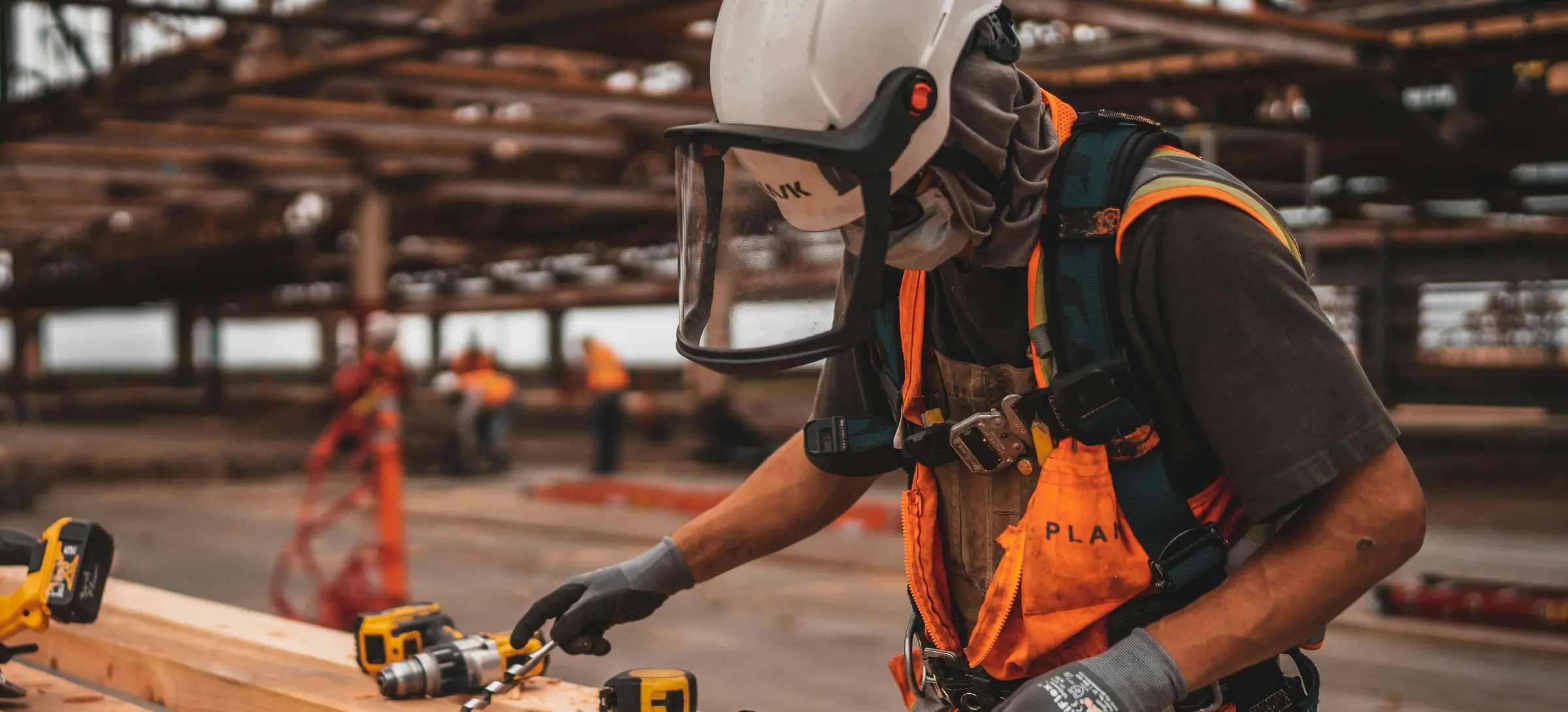 A worker in a helmet, visor and high-visibility harness drilling a pilot hole in a long timber beam in an open fabrication shed