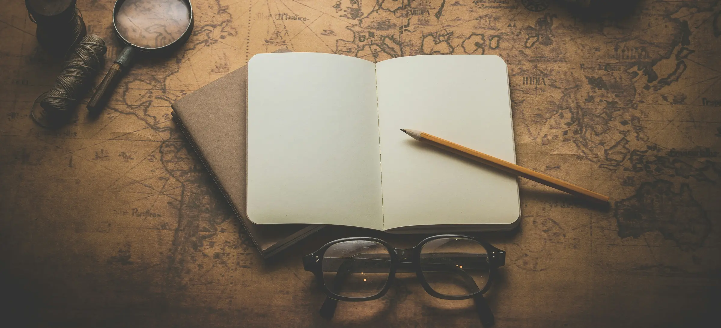 An open notebook with blank pages and a pencil, beside reading glasses and a magnifier on an antique map