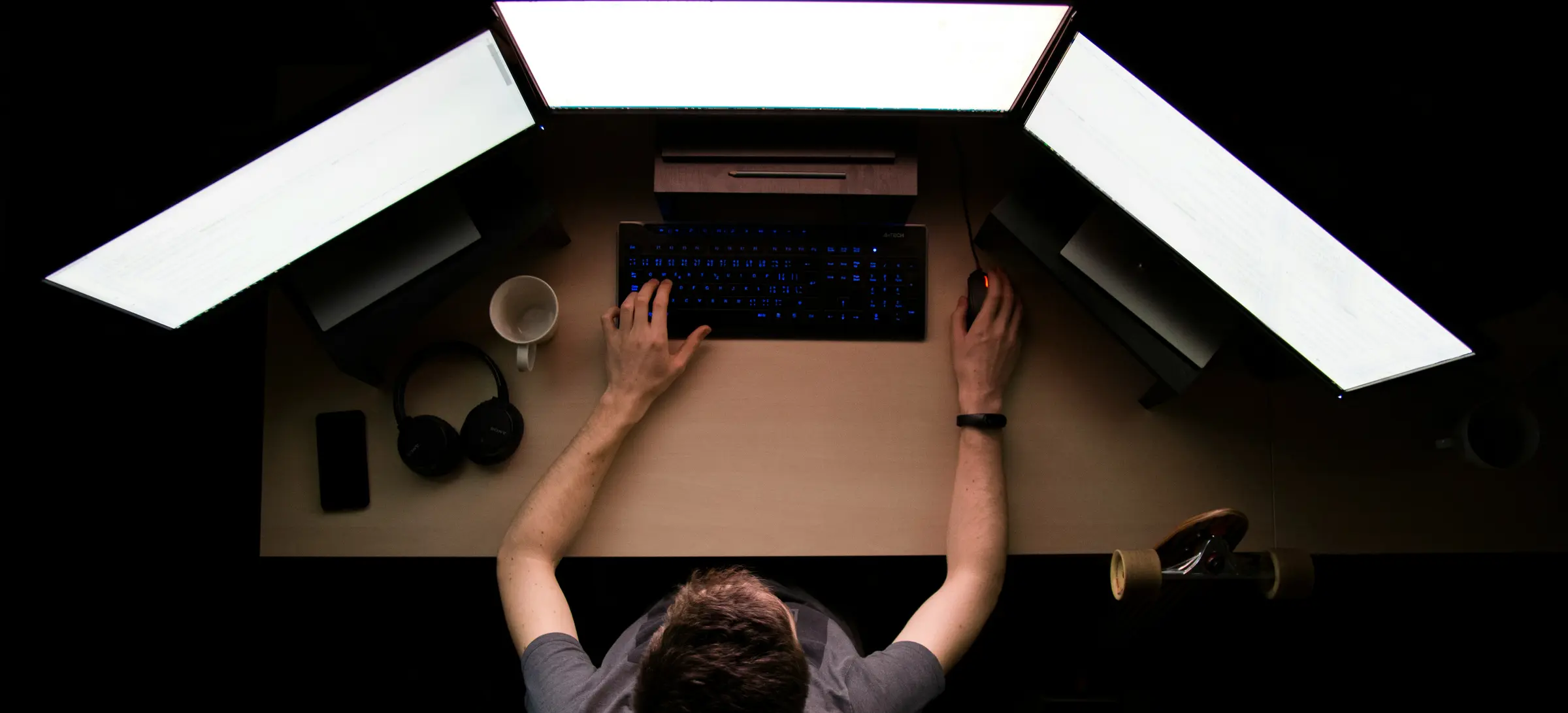 A desk seen from directly overhead in a dark room: three monitors, a lit keyboard, and a developer at work with both hands on the desk